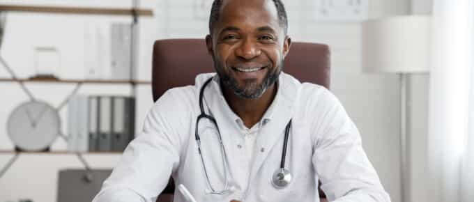 African American male doctor sitting in office chair at desk wearing white doctor coat and stethoscope around neck.