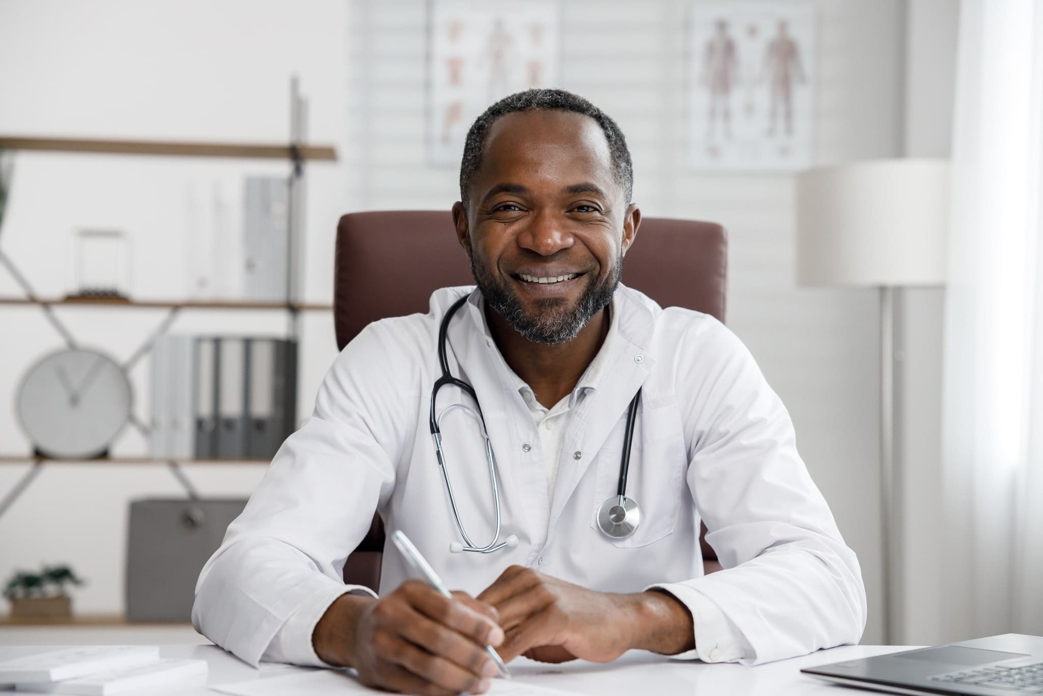 African American male doctor sitting in office chair at desk wearing white doctor coat and stethoscope around neck.