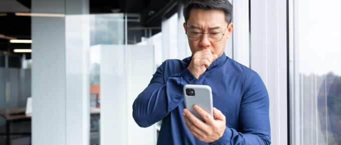 A worried and shocked young Asian man stands in front of large office window and looks at the phone in his hands.