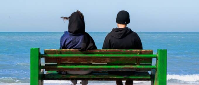 two adults sitting on a green bench looking out over blue water