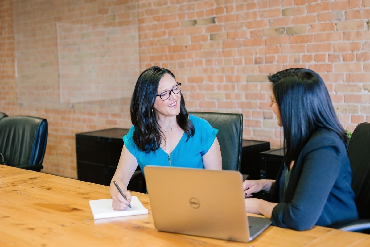 2 women with dark hair sitting at a desk with a laptop in front of a brick wall