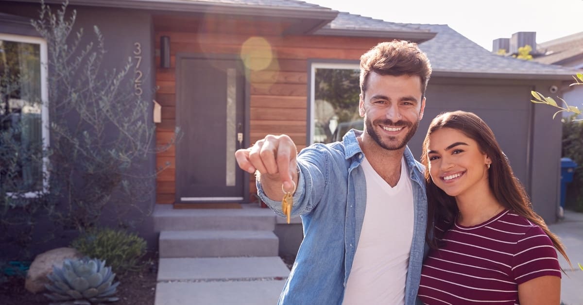 Portrait Of Couple Standing Outdoors In Front Of House With For Sale Sign In Garden Holding Keys