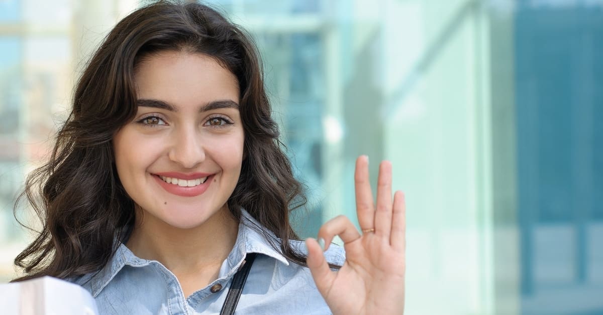 woman with dark hair wearing a blue shirt showing a sign that everything is OK with her hand.