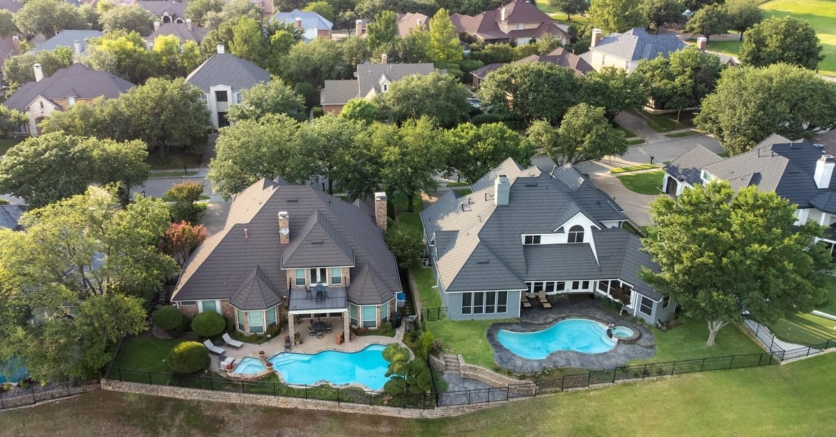 Aerial drone view of luxury mansions with swimming pools surrounded by green grass and trees in the summertime.