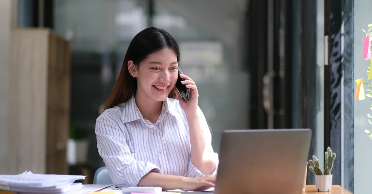 Portrait of young asian business woman talk on smartphone write notebook laptop in home office.