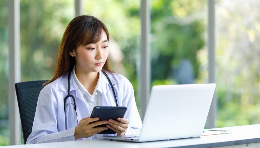 Asian successful professional young female doctor in white lab coat sitting at desk with laptop.