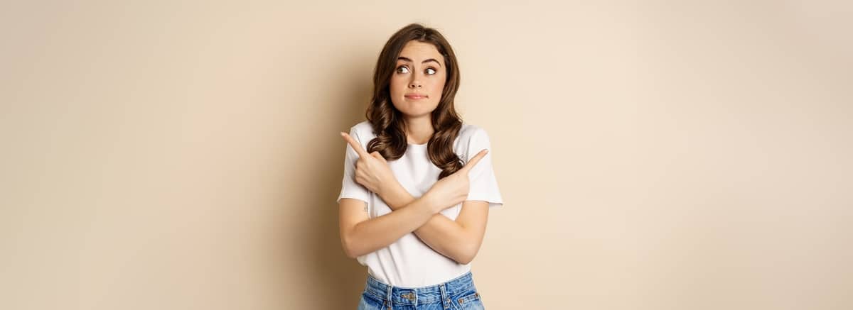 Girl choosing between two options, pointing sideways and looking clueless, standing over beige background
