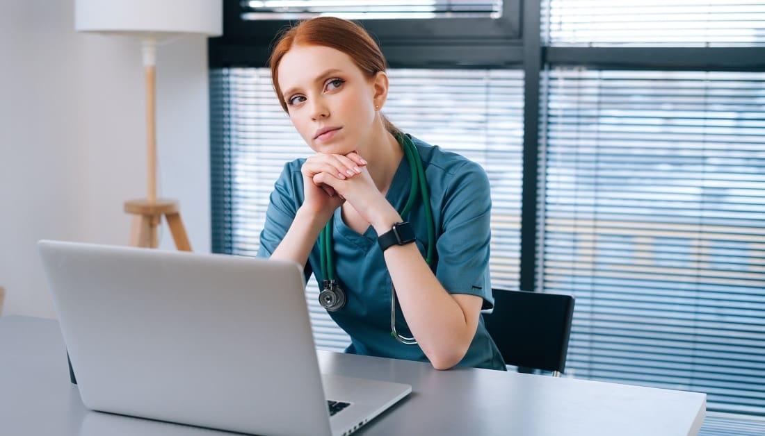 Portrait of thoughtful young female doctor in blue green medical uniform sitting at desk with laptop on background of window in hospital office of medic clinic, looking down.