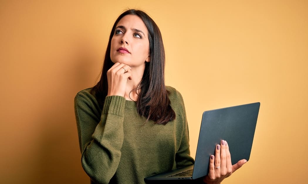 Young brunette woman with blue eyes working using computer laptop over yellow background with hand on chin thinking about question