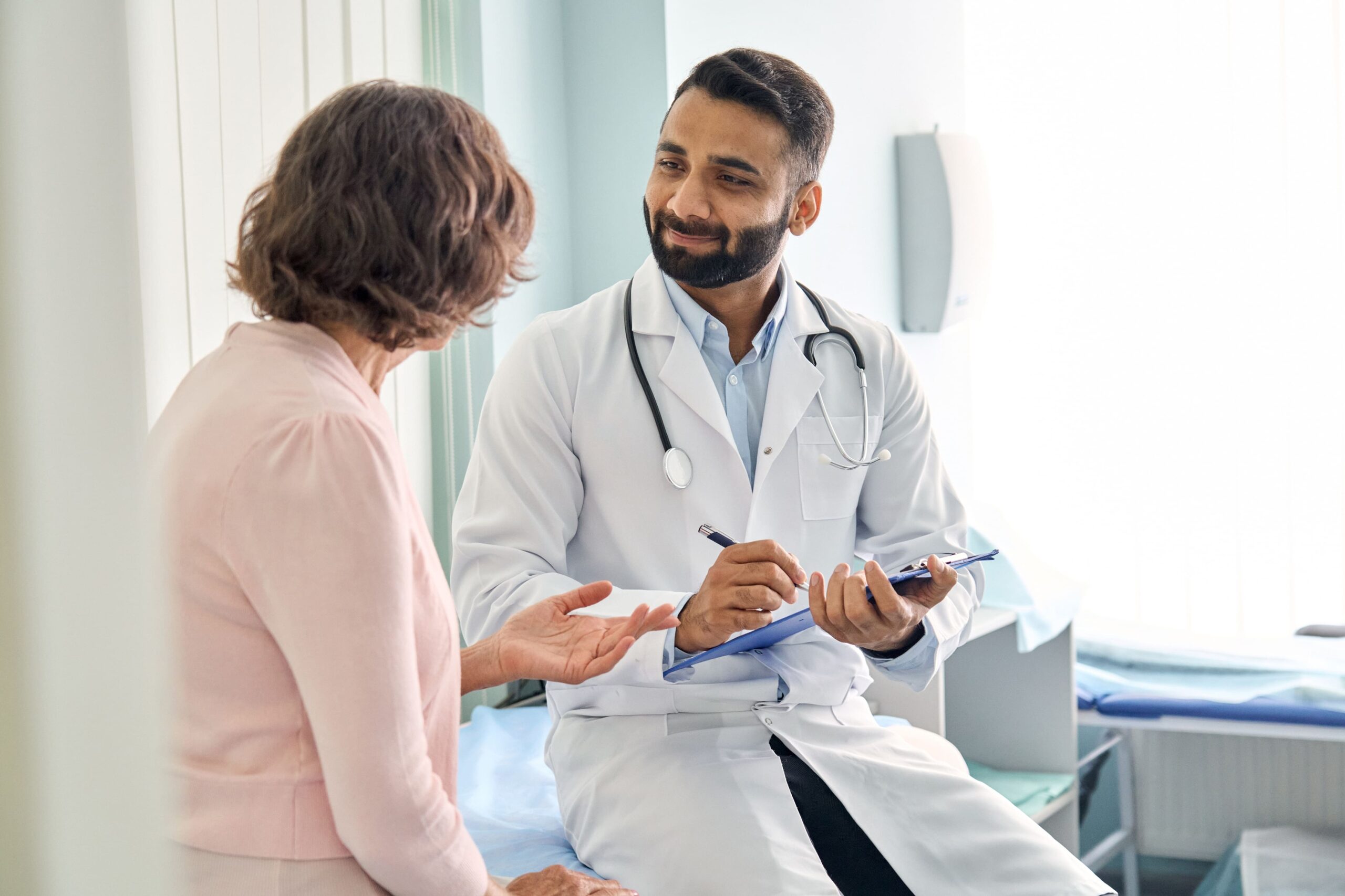 Male doctor with light brown skin and beard talking with an older female patient.