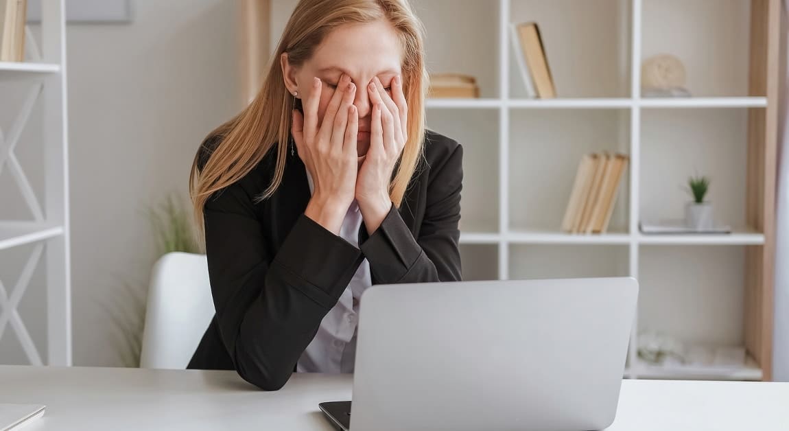 Stressed business woman sitting hands cover face in frustration at laptop at white office
