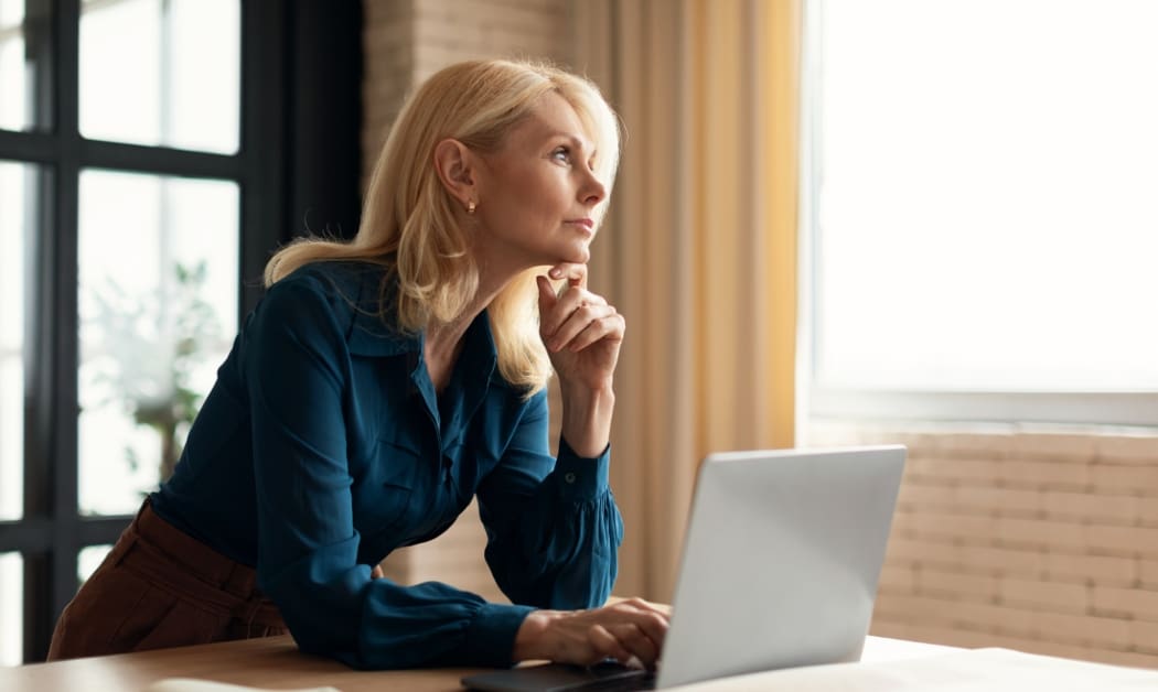 Woman with light colored hair sitting at a desk with a laptop looking out a window thoughtfully.