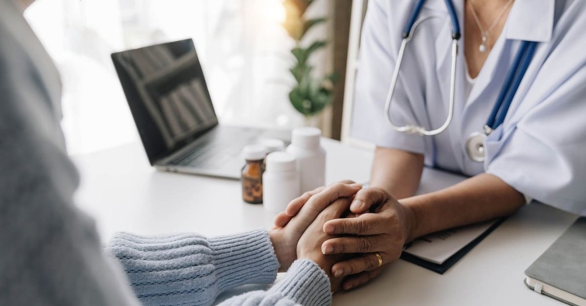 young woman nurse holding the hand of a senior woman sitting across from each other at a desk