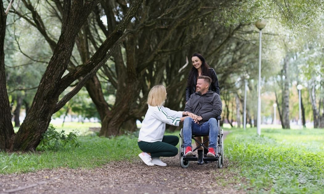 Happy disabled person in wheelchair and two women are talking to him in park.