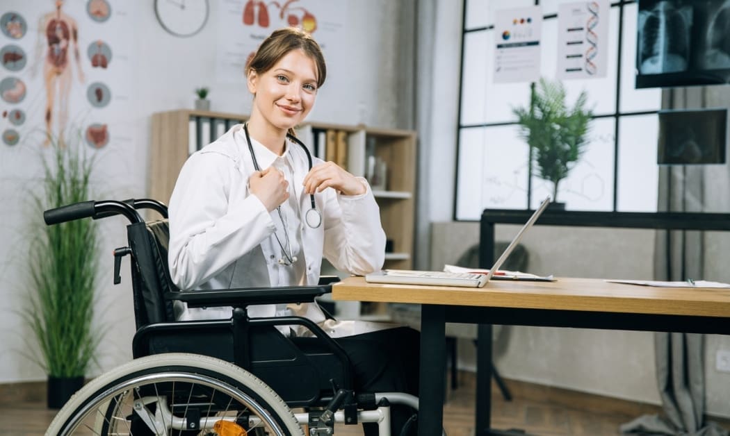 Female doctor in wheelchair smiling at camera while working on modern laptop at office room