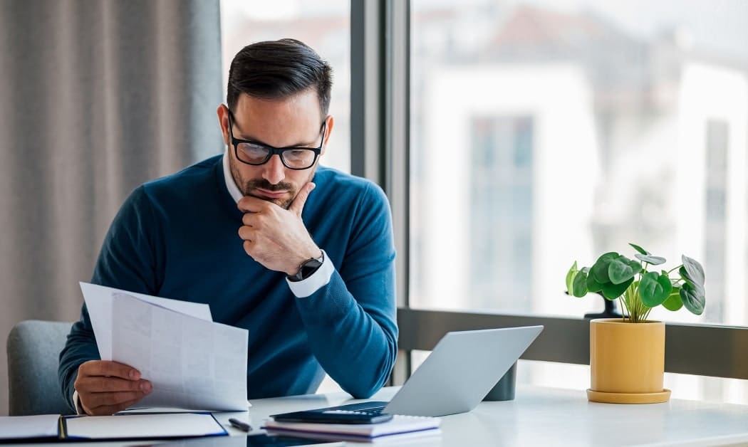 Dark haired man wearing a blue sweater sitting at a desk looking at a piece of paper in an office with a window