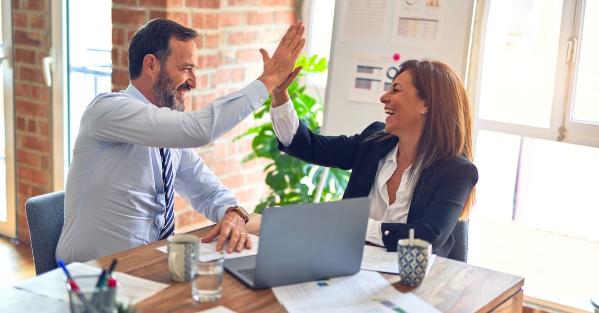 middle-aged man and woman high-fiving sitting at a conference table with a laptop and coffee