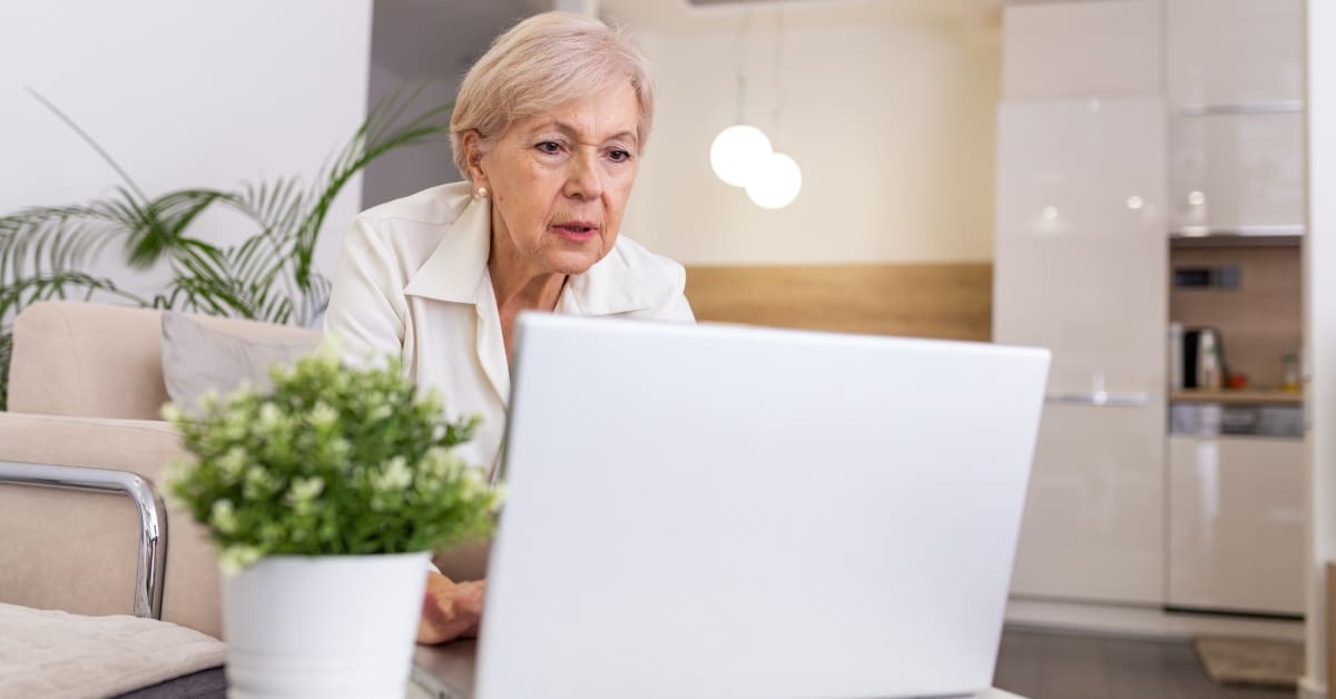 Senior woman sitting in a tan chair in front of a silver laptop on a table.