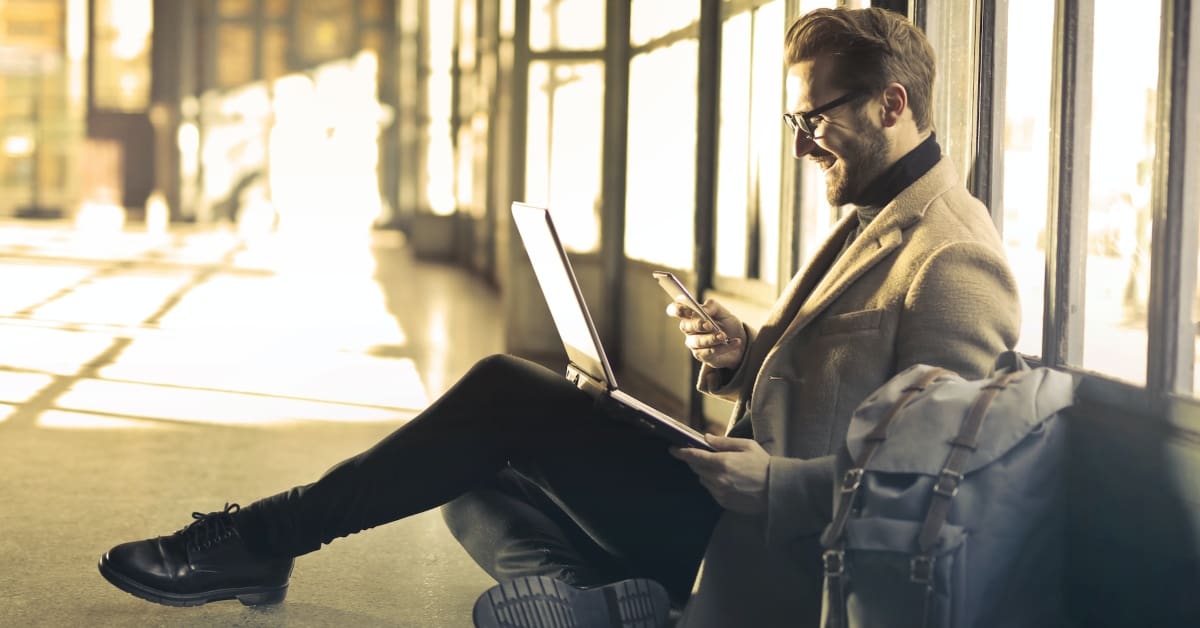 man wearing business suit leaning against a window with a laptop while looking at his cell phone