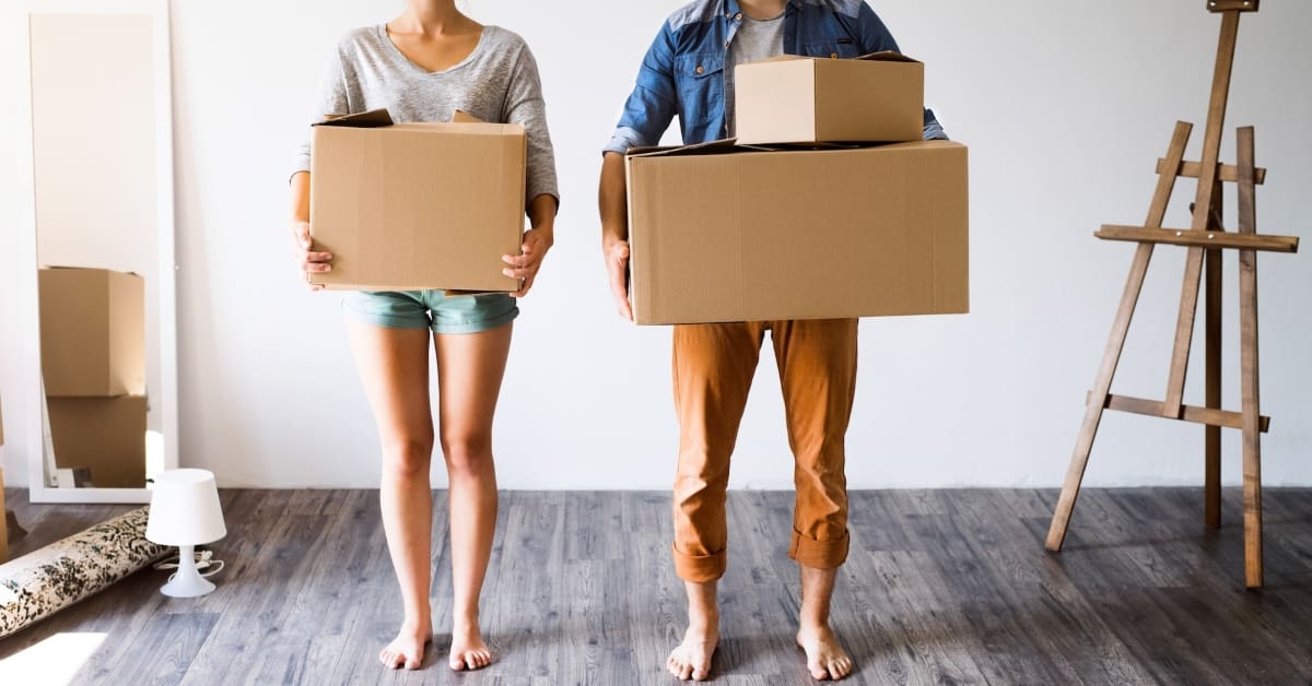 man and woman standing barefoot in an empty room, each holding a cardboard box.