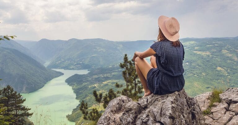 Woman in blue shirt wearing a tan hat sitting on a cliff top looking out over a river and green mountain landscape.