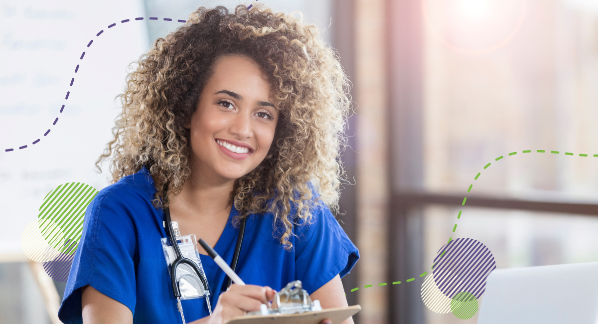 woman with brown hair wearing blue scrubs sitting at a desk while writing on a clipboard.