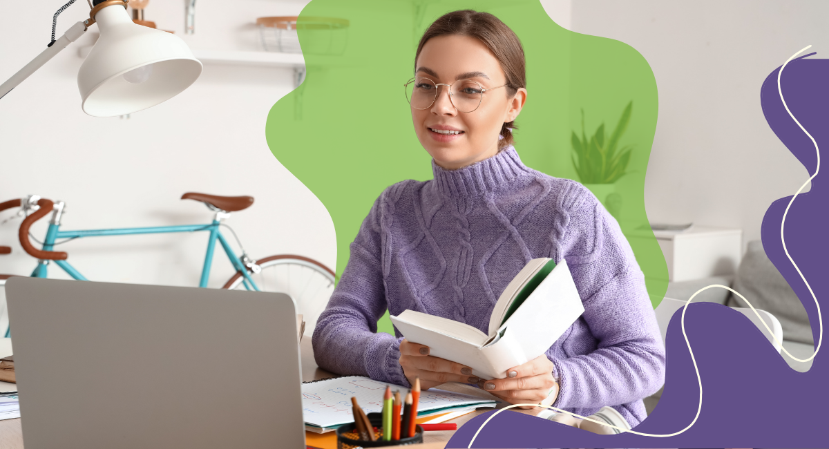 woman with brown hair sitting at a desk holding a book while looking at a laptop screen.