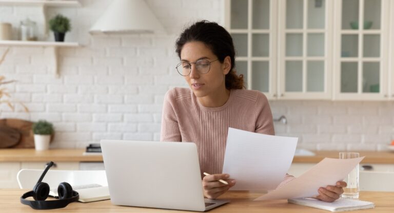 Female sitting at a home office desk, holding financial documents looking at a laptop