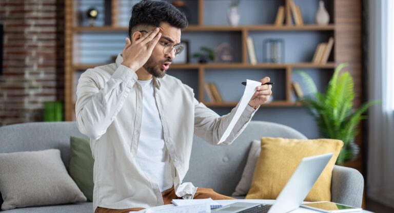Worried man holding receipt sitting on couch with laptop