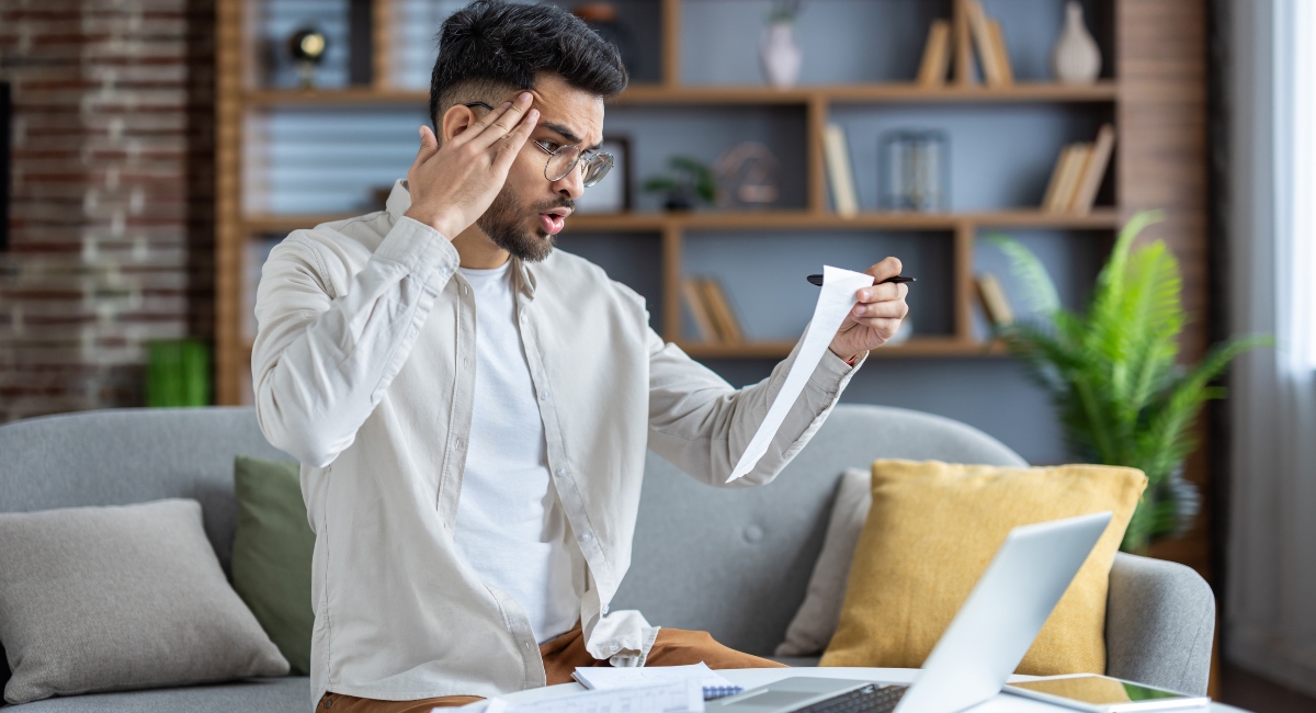 Worried man holding receipt sitting on couch with laptop